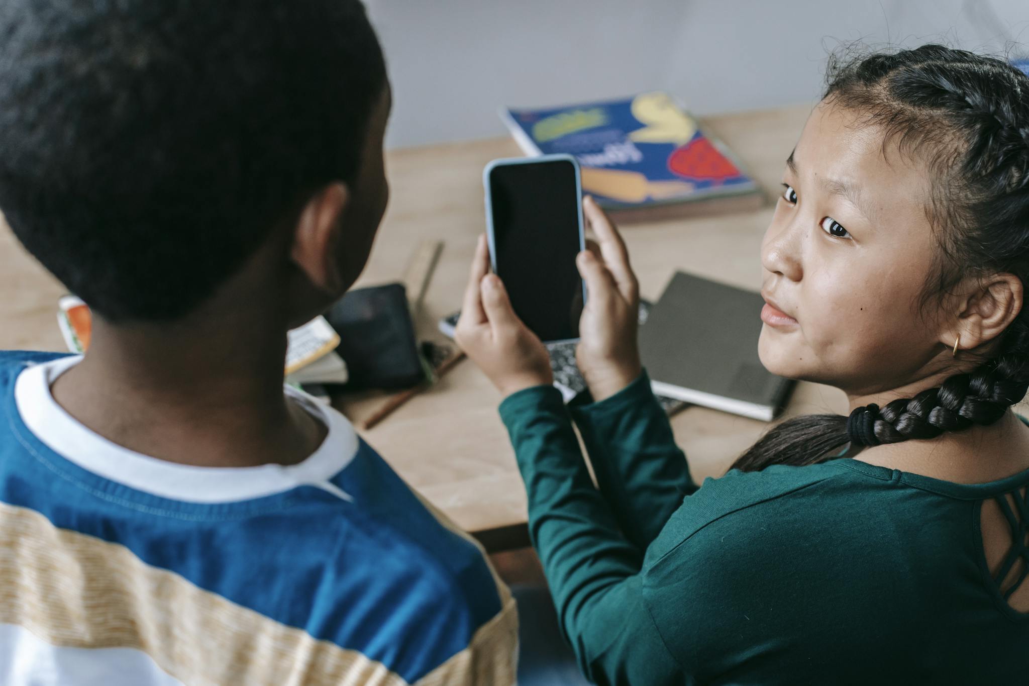 From above of adorable little Asian girl showing mobile phone to unrecognizable African American boy during break in classroom